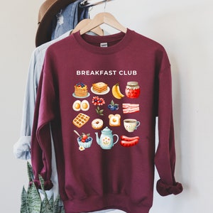 May include: A maroon sweatshirt with the text "BREAKFAST CLUB" printed in white. The sweatshirt features a graphic design of various breakfast foods, including pancakes, waffles, eggs, bacon, and coffee.