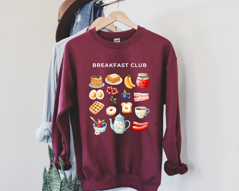 May include: A maroon sweatshirt with the text "BREAKFAST CLUB" printed in white. The sweatshirt features a graphic design of various breakfast foods, including pancakes, waffles, eggs, bacon, and coffee.