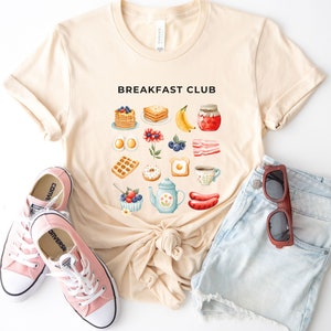May include: A beige t-shirt with the text "Breakfast Club" printed on the front. The shirt features a variety of breakfast foods, including waffles, pancakes, eggs, bacon, and coffee. The shirt is laid flat on a white surface with a pair of pink Converse shoes and a pair of blue denim shorts.