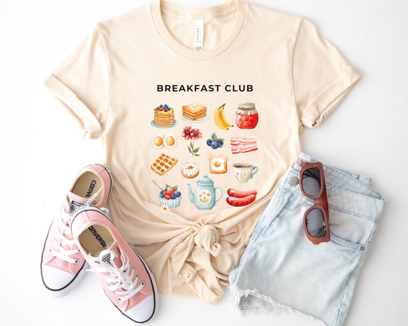 May include: A beige t-shirt with the text "Breakfast Club" printed on the front. The shirt features a variety of breakfast foods, including waffles, pancakes, eggs, bacon, and coffee. The shirt is laid flat on a white surface with a pair of pink Converse shoes and a pair of blue denim shorts.