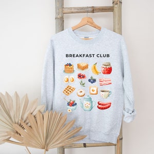 May include: A grey sweatshirt with the text "Breakfast Club" printed on the front. The sweatshirt features a variety of breakfast foods, including pancakes, waffles, eggs, bacon, and fruit.