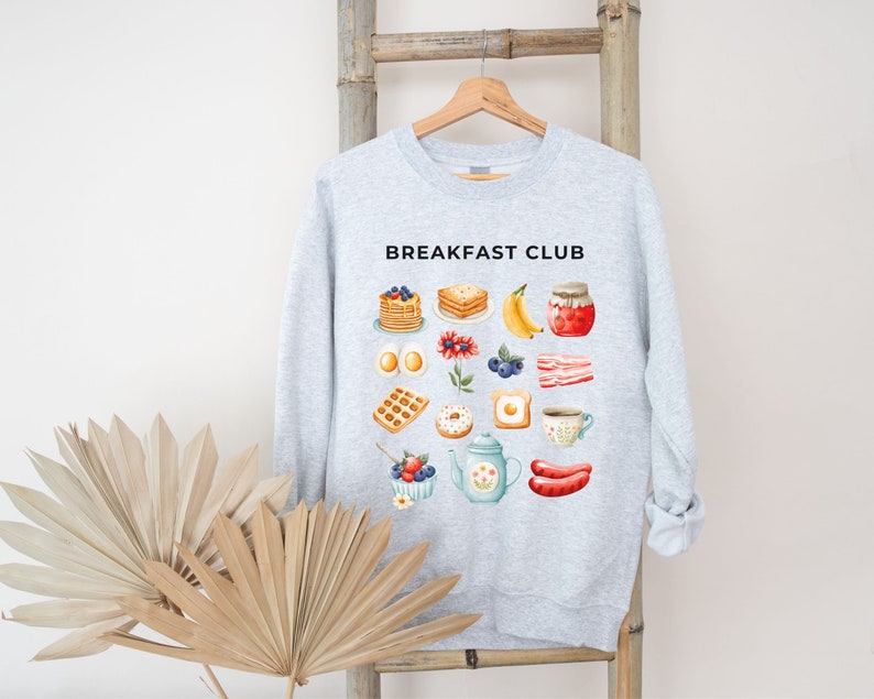 May include: A grey sweatshirt with the text "Breakfast Club" printed on the front. The sweatshirt features a variety of breakfast foods, including pancakes, waffles, eggs, bacon, and fruit.