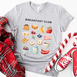 May include: A gray t-shirt with the text "Breakfast Club" printed on the front. The shirt features a variety of breakfast foods, including waffles, pancakes, eggs, bacon, and coffee.