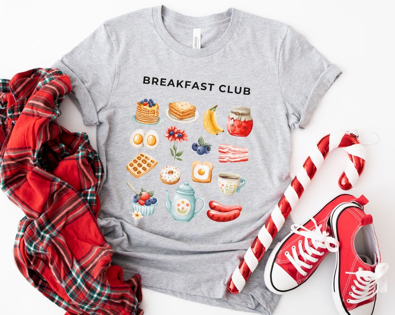 May include: A gray t-shirt with the text "Breakfast Club" printed on the front. The shirt features a variety of breakfast foods, including waffles, pancakes, eggs, bacon, and coffee.