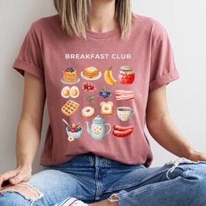 May include: A pink t-shirt with the text "Breakfast Club" printed on the front. The shirt features a variety of breakfast foods, including pancakes, waffles, eggs, bacon, and coffee.