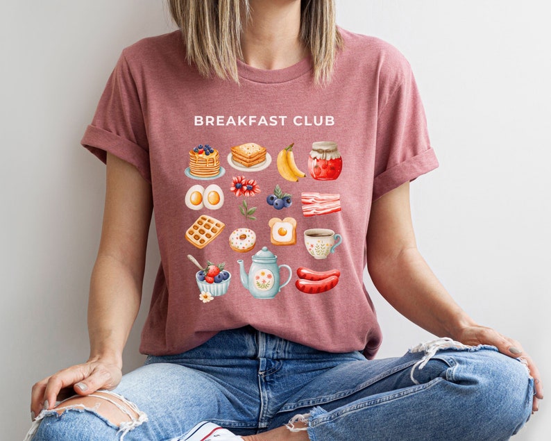 May include: A pink t-shirt with the text "Breakfast Club" printed on the front. The shirt features a variety of breakfast foods, including pancakes, waffles, eggs, bacon, and coffee.