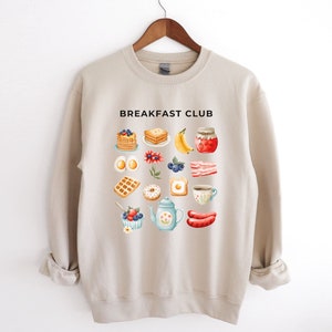 May include: Beige sweatshirt with the text "Breakfast Club" printed on the front. The sweatshirt features a graphic of various breakfast foods, including pancakes, eggs, bacon, waffles, doughnuts, and coffee.