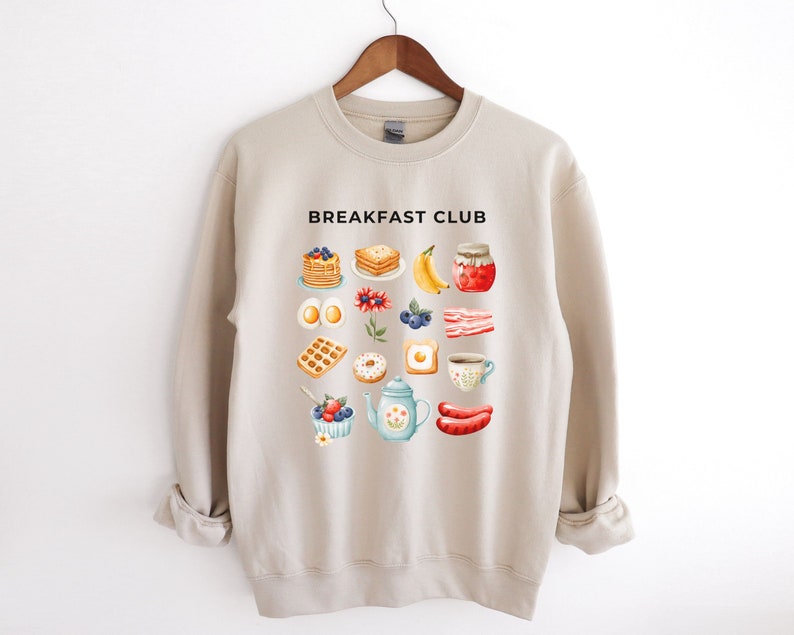 May include: Beige sweatshirt with the text "Breakfast Club" printed on the front. The sweatshirt features a graphic of various breakfast foods, including pancakes, eggs, bacon, waffles, doughnuts, and coffee.