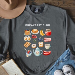 May include: A dark grey sweatshirt with the text "Breakfast Club" printed in white. The sweatshirt features a colourful illustration of various breakfast foods, including pancakes, waffles, eggs, bacon, and fruit.