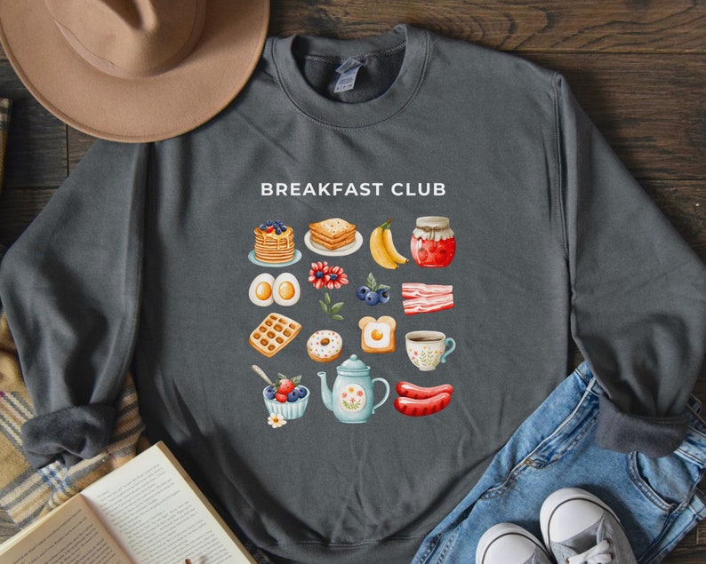 May include: A dark grey sweatshirt with the text "Breakfast Club" printed in white. The sweatshirt features a colourful illustration of various breakfast foods, including pancakes, waffles, eggs, bacon, and fruit.