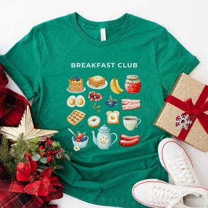 May include: Green t-shirt with the text "Breakfast Club" printed on the front. The shirt features a grid of colorful illustrations of breakfast foods, including waffles, eggs, bacon, fruit, and coffee.