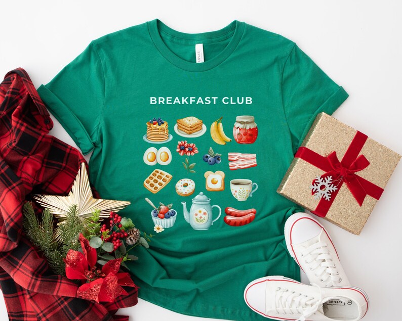 May include: Green t-shirt with the text "Breakfast Club" printed on the front. The shirt features a grid of colorful illustrations of breakfast foods, including waffles, eggs, bacon, fruit, and coffee.