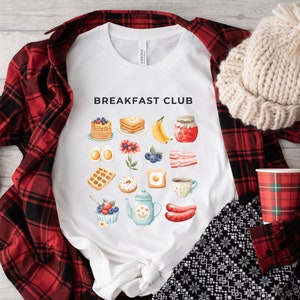 May include: White t-shirt with the text "Breakfast Club" printed on the front. The design features a variety of breakfast foods, including pancakes, eggs, bacon, and coffee.