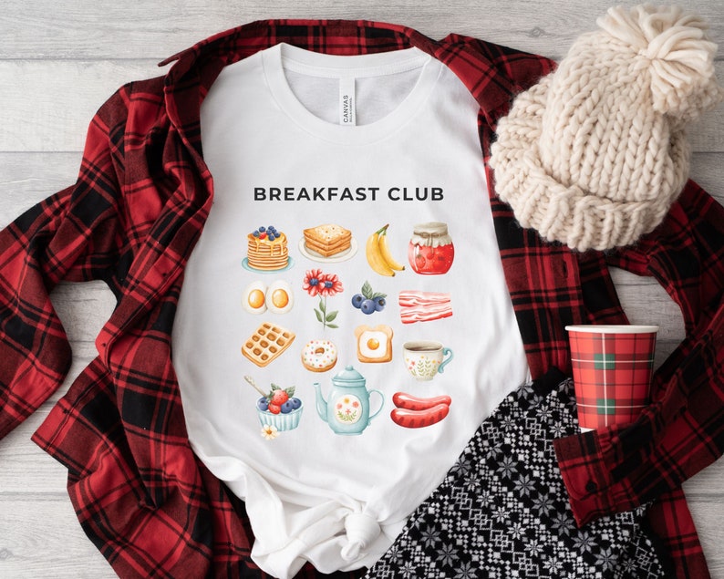 May include: White t-shirt with the text "Breakfast Club" printed on the front. The design features a variety of breakfast foods, including pancakes, eggs, bacon, and coffee.