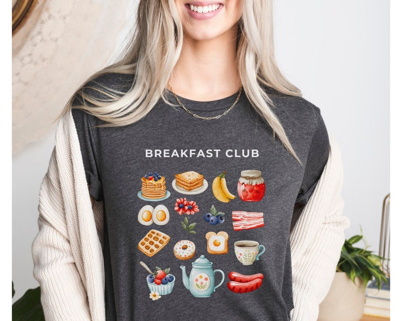 May include: A dark gray t-shirt with the text "Breakfast Club" printed on the front. The shirt features a colorful illustration of breakfast foods, including waffles, eggs, bacon, pancakes, and coffee.