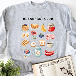 May include: Grey sweatshirt with the text "BREAKFAST CLUB" printed in black. The sweatshirt features a colourful illustration of various breakfast foods, including pancakes, waffles, eggs, bacon, doughnuts, and coffee.