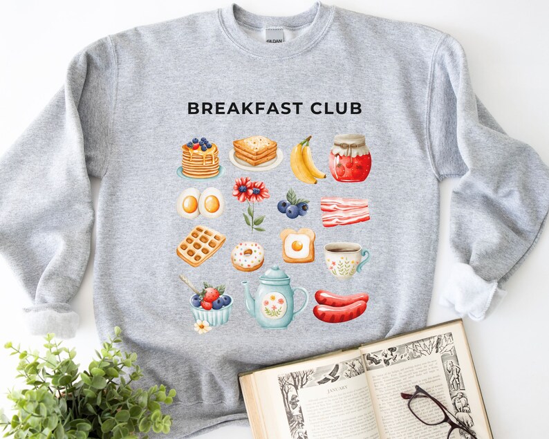 May include: Grey sweatshirt with the text "BREAKFAST CLUB" printed in black. The sweatshirt features a colourful illustration of various breakfast foods, including pancakes, waffles, eggs, bacon, doughnuts, and coffee.