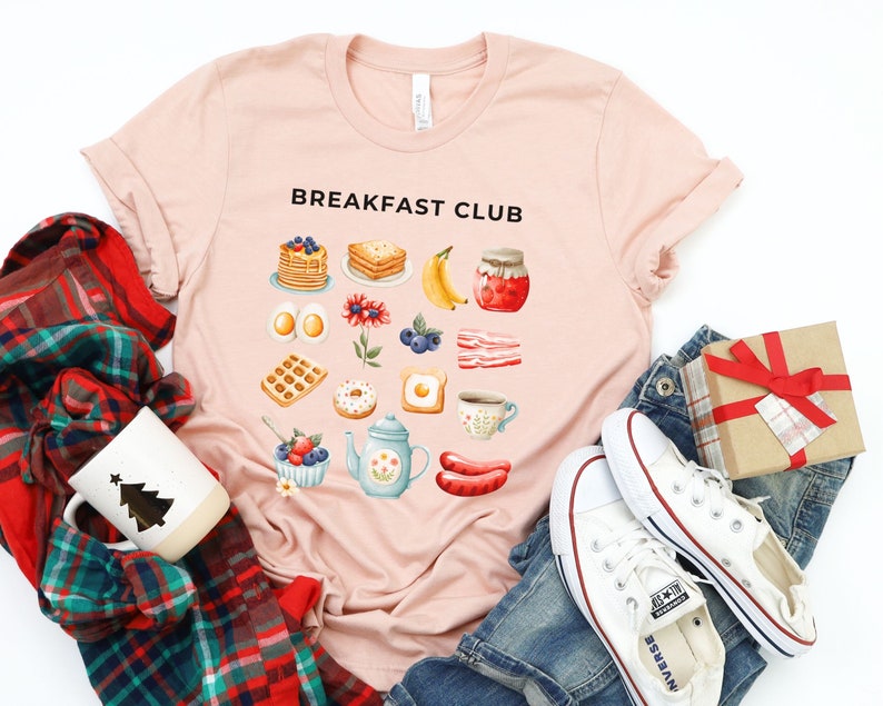 May include: A pink t-shirt with the text "Breakfast Club" printed on the front. The shirt features a graphic design of various breakfast foods, including waffles, eggs, bacon, donuts, and coffee.
