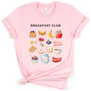 May include: Pink t-shirt with the text "Breakfast Club" and a graphic of various breakfast foods, including pancakes, eggs, bacon, waffles, donuts, coffee, and fruit.