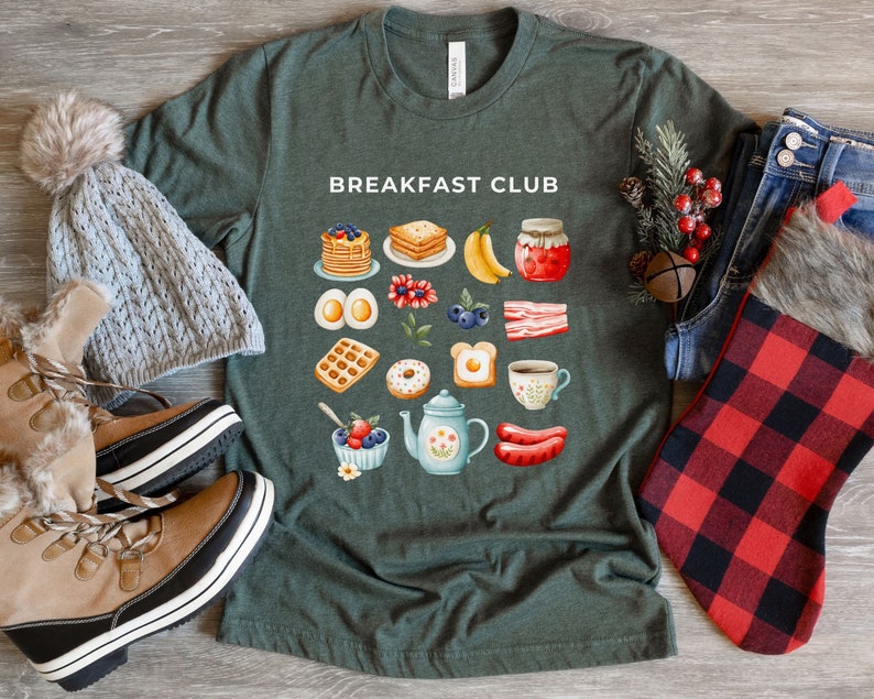 May include: A green t-shirt with the text "Breakfast Club" printed on the front. The shirt features a variety of breakfast foods, including waffles, eggs, bacon, and coffee.