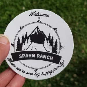 May include: A round white sticker with the text "Welcome to Spahn Ranch" in a black compass design. The sticker features a mountain, trees, and a tent graphic. The text "Where we're one big happy family!" is also included.