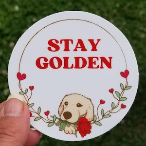 以下が含まれることがあります： 赤い文字で「STAY GOLDEN」と書かれた丸い白いステッカー。赤いバラを持った漫画のゴールデンレトリバーが下にいます。デザインは茶色の線とハートと葉のガーランドで縁取られています。