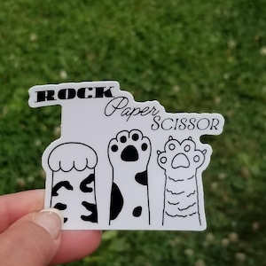 May include: White sticker with black text and paw print illustrations. The text reads "ROCK Paper SCISSOR".