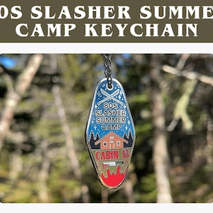 May include: A metal keychain with the text "80s Slasher Summer Camp Keychain". The keychain has a blue background with a cabin, crossed knives, and the words "Cabin 13". The bottom of the keychain has a red blood-like design.