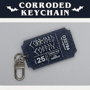 May include: A black and silver keychain shaped like a ticket stub with the text "Corroded Coffin" and "032786 Gen. Admission" printed on it. The ticket stub is decorated with glitter.