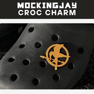 May include: A gold Mockingjay charm featuring a bird and arrow design. The charm is attached to a black shoe.