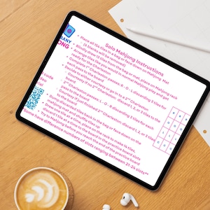 May include: A tablet displaying Solo Mahjong Instructions, with text detailing game setup and gameplay. The tablet is on a wooden surface, accompanied by a cup of coffee and wireless earbuds. The instructions include steps for the Charleston and discarding tiles.