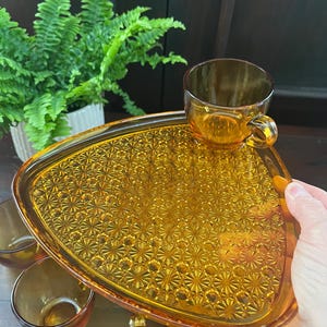 Puede incluir: Un juego de tazas de vidrio ámbar y una bandeja de servir triangular a juego con un patrón de estrella. La bandeja tiene un asa en un lado.