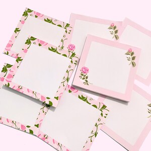 May include: A collection of square notepads with a pink border and a floral design. The border features pink flowers and green leaves. A pink rose is printed in the corner of some notepads. The notepads have a white interior.