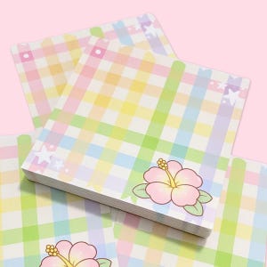 Può includere: Una pila di blocchi per appunti quadrati dai colori pastello con un motivo a quadretti nei toni del rosa, giallo, verde e blu. Ogni blocco per appunti presenta un'illustrazione di fiore di ibisco rosa con foglie verdi. Gli angoli superiori hanno disegni a stella e a pois.