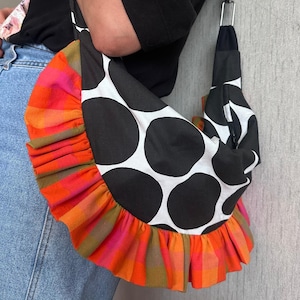 Puede incluir: Un bolso de hombro estilo hobo con un patrón de lunares grandes en blanco y negro en el cuerpo principal, acentuado por un volante a cuadros naranja, rosa y verde vibrante a lo largo del borde curvo, unido con una correa de metal plateado.