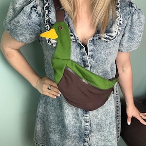 May include: A crossbody bag shaped like a duck, featuring a green head and neck, a yellow beak, a brown body, and a brown adjustable strap. The bag includes a metal zipper closure across the middle of the duck's body and a small brown button eye.