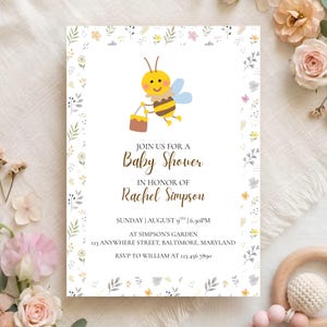May include: A rectangular baby shower invitation card featuring a cartoon bee carrying a honey pot, surrounded by a floral border. The text reads: JOIN US FOR A Baby Shower IN HONOR OF RACHEL SIMPSON, SUNDAY | AUGUST 9TH | 6:30PM, AT SIMPSON'S GARDEN.