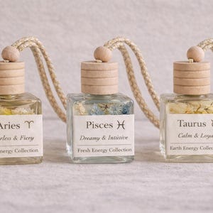May include: Three square glass bottles with wooden caps and beige cords. Each bottle contains dried flowers and a label with zodiac signs: Aries, Pisces, and Taurus. The labels also include the phrases "Fearless & Fiery", "Dreamy & Intuitive", and "Calm & Loyal".