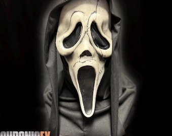 May include: A white, elongated ghost mask with black eye sockets and a black mouth opening, featuring dark crack-like detailing across the surface. The mask is attached to a black fabric hood that drapes down the sides and covers the neck area.