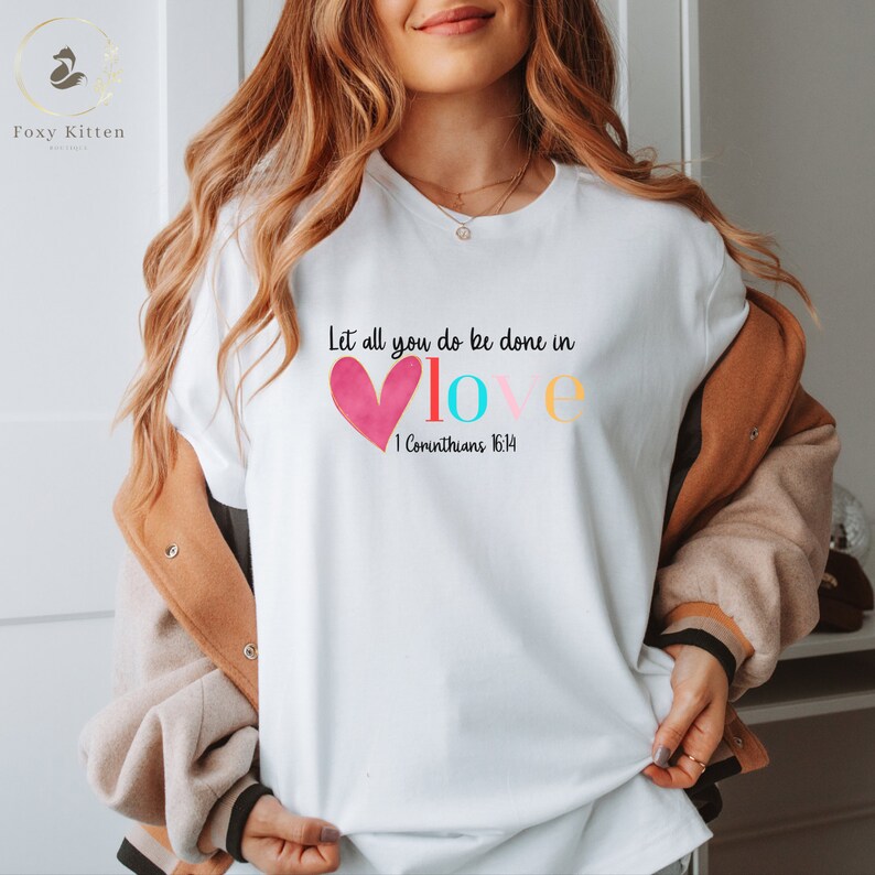 May include: White t-shirt with a pink heart and the words "Let all you do be done in love" in colorful letters. The text "1 Corinthians 16:14" is below the heart.