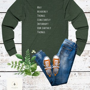 以下が含まれることがあります： 前面に「May Heavenly Things Constantly Interrupt Our Earthly Things」という文字がプリントされた、長袖のフォレストグリーンのTシャツ。このTシャツは、青いジーンズとオレンジのスニーカーと組み合わせています。