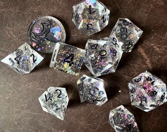 May include: A set of nine iridescent dice with black numbers. The dice are clear with a glittery finish and are arranged on a brown surface. The dice are numbered 1 through 6 and include a d20, d12, d10, d8, d6, and d4.