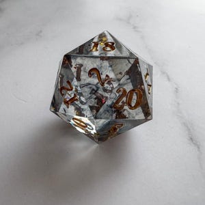 May include: A clear, twenty-sided die with gold-colored numbers and internal swirls of gray and brown. The die is designed for tabletop role-playing games.