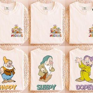 May include: Cream-colored t-shirts featuring cartoon characters and names. The top row displays a group of characters with the name "NICOLAS" and "JESSICA". The bottom row shows individual characters with the names "HAPPY", "SLEEPY", and "DOPEY".