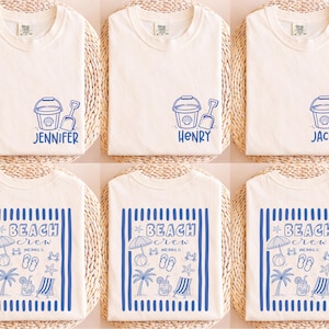 May include: Cream-colored t-shirts with blue graphics. Some shirts feature a bucket and shovel design with names like "Jennifer", "Henry", and "Jackson". Other shirts display "Beach Crew" text with beach-themed illustrations, including an umbrella, palm trees, and a beach chair.