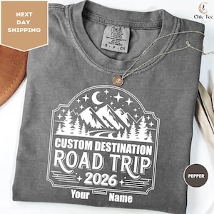 a gray t-shirt with a white design featuring a mountain landscape and stars, along with the text "Custom Destination Road Trip 2026 Your Name". The t-shirt is displayed on a white background, and there is a small text box in the top left corner that reads "Next day shipping".