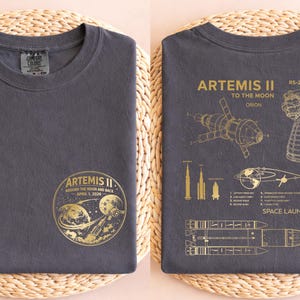 May include: Dark gray t-shirt featuring a gold graphic of the Artemis II mission, including a rocket, the moon, and the text "ARTEMIS II Around the Moon and Back April 1, 2020." The back of the shirt displays diagrams and text related to the mission.