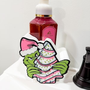 Puede incluir: Soporte decorativo para dispensador de jabón con un personaje de dibujos animados verde que lleva un gorro de Papá Noel rosa y sostiene un árbol de Navidad. El soporte es blanco y está diseñado para contener una botella de jabón.