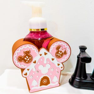 Könnte beinhalten: Ein rosa-brauner Seifenspender mit Lebkuchenhaus-Design und Mickey Mouse-Details. Der Spender hat eine weiße Pumpe und eine transparente Flasche mit rosa Flüssigkeit. Das Design umfasst Mickey Mouse-Silhouetten und eine Lebkuchenhausform.