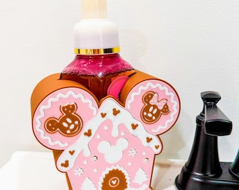 May include: A pink and brown soap dispenser featuring a gingerbread house design with Mickey Mouse details. The dispenser has a white pump and a clear bottle filled with pink liquid. The design includes Mickey Mouse silhouettes and a gingerbread house shape.